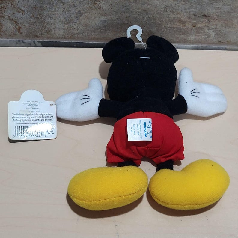 K&ouml;nnte beinhalten: Ein Pl&uuml;schtier von Mickey Mouse, von hinten gesehen, mit schwarzem Kopf und K&ouml;rper, roter Hose, wei&szlig;en Handschuhen und gelben Schuhen. Das Spielzeug hat ein Anh&auml;ngeetikett. Das Spielzeug wird von Applause hergestellt.