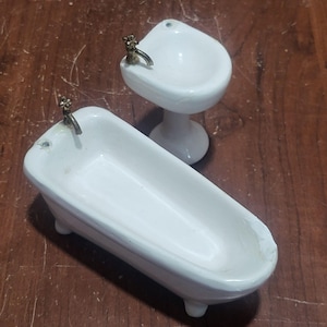 May include: A miniature white porcelain bathtub and pedestal sink set. The bathtub has four small feet and a brass faucet. The sink has a brass faucet and a pedestal base. The set is designed for a dollhouse or miniature display.