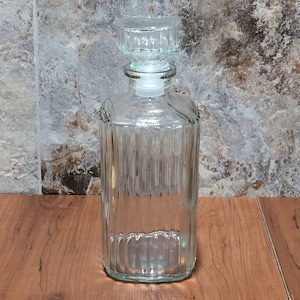 May include: A clear glass bottle with a textured surface and a glass stopper. The bottle is rectangular in shape and is empty.