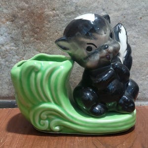 May include: A green ceramic planter shaped like a leaf with a black and white skunk figurine sitting on top. The skunk is holding its tail up and has a playful expression.