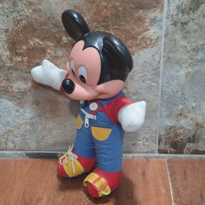 May include: A Mickey Mouse plush toy, featuring a black head and ears, a pink face, and white gloves. He wears blue overalls with yellow accents, a red shirt, and red and yellow shoes. The toy is standing on a brown surface.