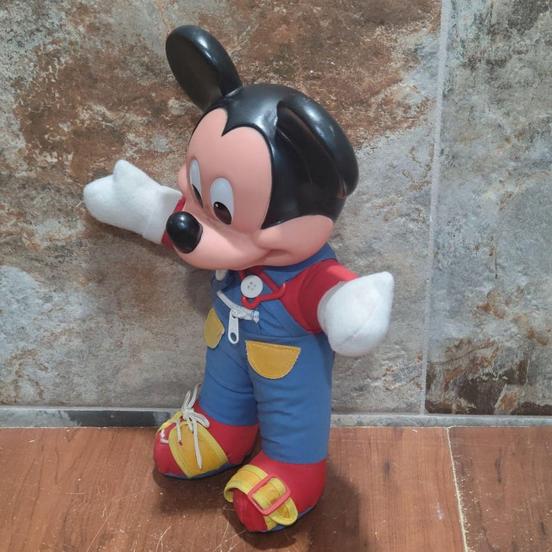 May include: A Mickey Mouse plush toy, featuring a black head and ears, a pink face, and white gloves. He wears blue overalls with yellow accents, a red shirt, and red and yellow shoes. The toy is standing on a brown surface.