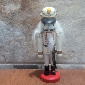 Puede incluir: Una figura de cascanueces de madera con uniforme militar. La figura tiene un abrigo blanco con detalles en negro y dorado, barba blanca y sombrero con emblema dorado. Se apoya sobre una base circular roja. Decoración navideña clásica.