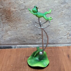 May include: A green frog figurine perched on a metal stand, with a smaller green frog figurine sitting on a green lily pad base.