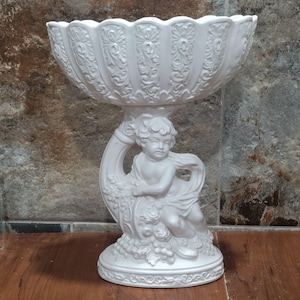 Könnte beinhalten: Weiße Keramikschale mit einem Cherub-Design. Die Schale steht auf einem Sockel mit einem Cherub, der Trauben hält. Die Schale ist verziert und hat einen gewellten Rand.