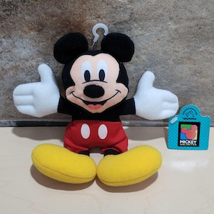 K&ouml;nnte beinhalten: Ein Mickey Mouse Pl&uuml;schtier mit schwarzen Ohren, einem schwarzen K&ouml;rper, roten Shorts und gelben Schuhen. Das Spielzeug hat wei&szlig;e Handschuhe und l&auml;chelt mit offenen Armen. Eine kleine Karte mit dem Text "Mickey Unlimited" befindet sich neben dem Spielzeug.
