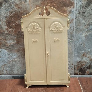May include: A beige plastic toy wardrobe with two doors, featuring a decorative arched top and hinges on the sides. The doors are embossed with the text 'Susy Goose' and 'Barbie' in a raised font, with a small central handle on the right door.