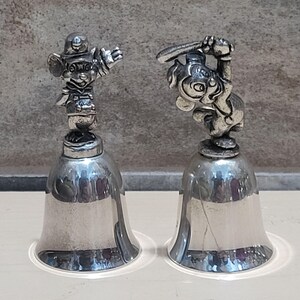 Pu&ograve; includere: Due campane color argento con figurine di personaggi dei cartoni animati in cima. Una campana presenta un personaggio in uniforme da football, l'altra tiene una mazza da baseball. Le campane hanno una forma classica.