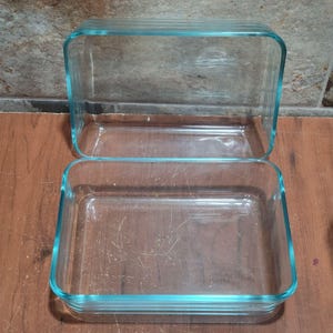 May include: Two clear glass rectangular baking dishes with a light blue trim. The dishes are empty and appear to be clean. The dishes are on a wooden surface.