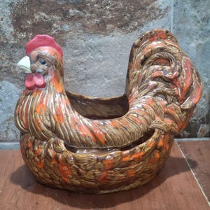 May include: A ceramic rooster-shaped planter with a brown, orange, and red color scheme. The planter has a detailed design, including a red comb and wattle, and a curved tail. The planter is designed to hold plants or other decorative items.