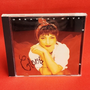 May include: A CD case featuring a portrait of Gloria Estefan. The album cover has a red background with the words "Greatest Hits" at the top. The artist's name, "Gloria," is written in a stylized script. The image is set against a red background.