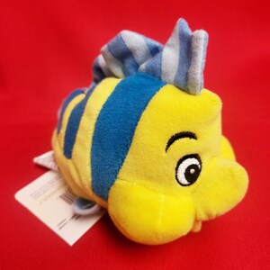 K&ouml;nnte beinhalten: Ein gelbes Pl&uuml;schfischspielzeug mit blauen Streifen und einer blau-wei&szlig; gestreiften Flosse. Der Fisch hat ein l&auml;chelndes Gesicht mit einem schwarzen Auge und einer wei&szlig;en Pupille. Ein wei&szlig;es Etikett ist am Fisch befestigt.