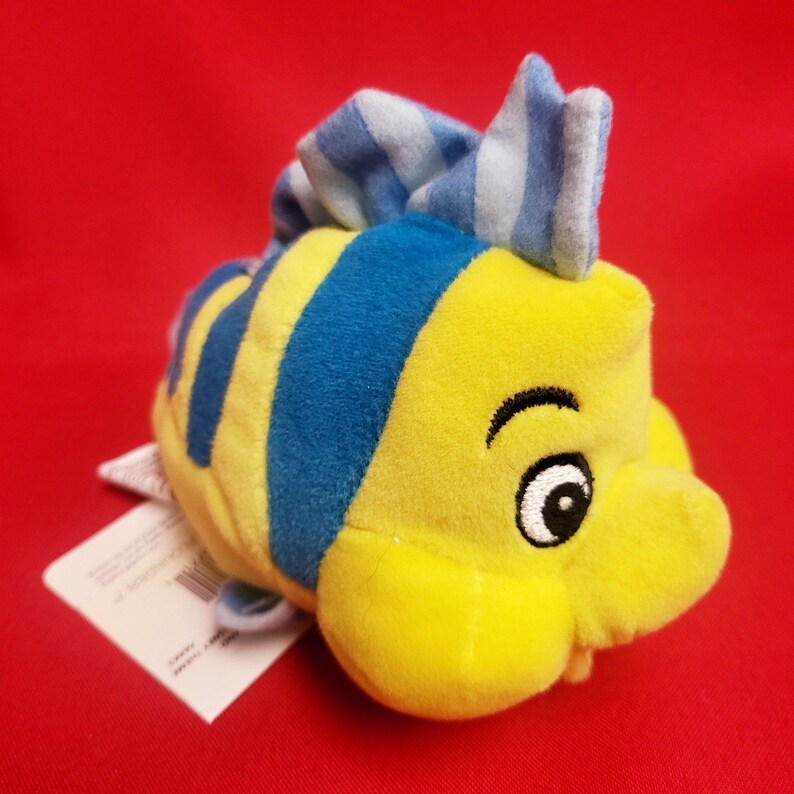 K&ouml;nnte beinhalten: Ein gelbes Pl&uuml;schfischspielzeug mit blauen Streifen und einer blau-wei&szlig; gestreiften Flosse. Der Fisch hat ein l&auml;chelndes Gesicht mit einem schwarzen Auge und einer wei&szlig;en Pupille. Ein wei&szlig;es Etikett ist am Fisch befestigt.