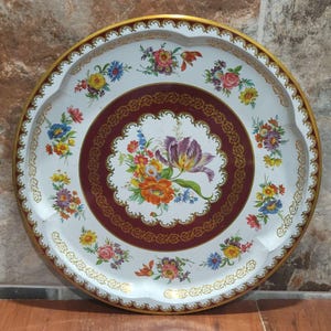 Puede incluir: Una bandeja redonda de metal blanco con borde festoneado y borde dorado. La bandeja presenta un diseño floral con un centro burdeos y un borde decorativo dorado. Las flores son en tonos morado, naranja, amarillo, rosa y azul.