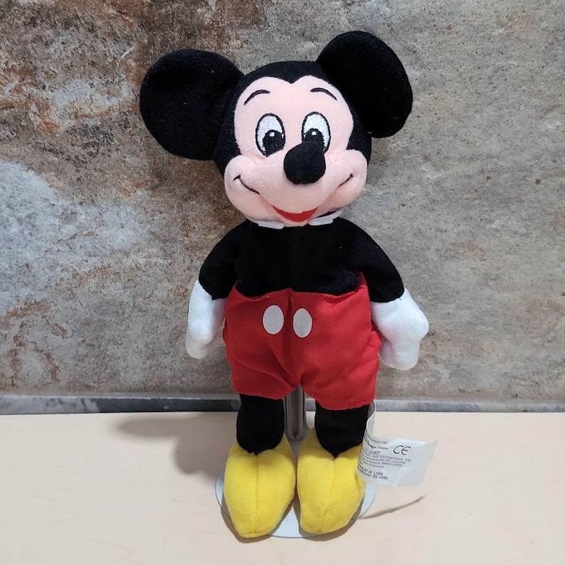 May include: A Mickey Mouse plush toy. The plush has a black head with large ears, a pink face, and a black nose. It wears a black shirt, red shorts with white buttons, white gloves, and yellow shoes. A white tag is attached to the toy.