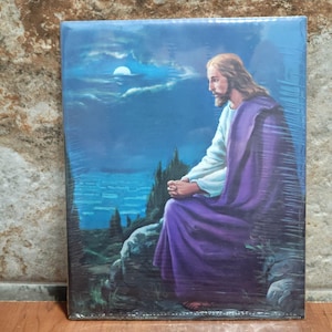 May include: A religious art print depicting a figure with long hair and a beard, wearing a white robe and a purple cloak, seated on a rock formation. The background features a night sky with a full moon and a dark landscape.