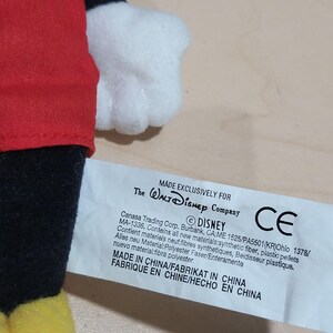 May include: A close-up of a Mickey Mouse plush toy with a red shirt, black shorts, and yellow shoes. The white glove is visible. A tag reads "MADE EXCLUSIVELY FOR The Walt Disney Company" and "DISNEY".