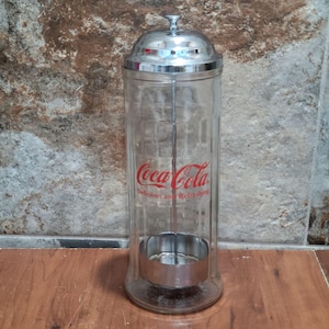 May include: A vintage Coca-Cola syrup dispenser with a clear glass body and a shiny chrome lid and base. The Coca-Cola logo is printed in red on the front, with the words "Delicious and Refreshing" below. The dispenser is designed to hold syrup for making soda.