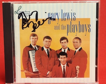 Puede incluir: Caja de CD con la portada del álbum "Gary Lewis and the Playboys". La portada tiene un fondo amarillo con el nombre de la banda en azul y una foto de los miembros de la banda con chaquetas rojas. La caja del CD está firmada.
