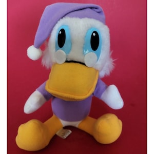 May include: Purple and white plush toy duck wearing a purple sleep cap. The duck has large blue eyes and a yellow beak.