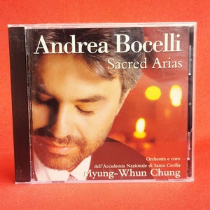May include: A CD case featuring Andrea Bocelli's album "Sacred Arias." The cover displays a close-up of Bocelli's face with the album title in white text. The orchestra and chorus are credited at the bottom.