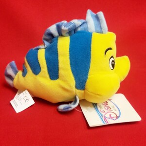K&ouml;nnte beinhalten: Ein Pl&uuml;schtier der Figur Fabius aus Disneys Arielle, die Meerjungfrau. Der gelb-blau gestreifte Fisch hat ein l&auml;chelndes Gesicht und eine blaue Flosse. Das Spielzeug hat ein Etikett von The Disney Store.