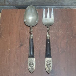 Könnte beinhalten: Ein Set aus einem silberfarbenen Metalllöffel und einer dreizinkigen Gabel. Jedes Besteckteil hat einen gedrehten Metallhals, einen glatten schwarzen Griff mit zwei kleinen Nieten und eine dekorative Endkappe mit einer sitzenden Figur.
