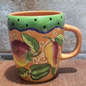 May include: A ceramic mug with a vibrant design featuring colorful vegetables. The mug has an orange base with painted illustrations of eggplants, peppers, and leaves. The rim is green with black dots, and the handle is orange.