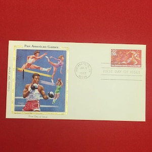 May include: A cream-colored first day of issue envelope featuring a Pan American Games cachet with illustrations of athletes, a 22-cent USA postage stamp, and a postmark dated Jan 29, 1987, from Indianapolis, IN, with the text Pan American Games.