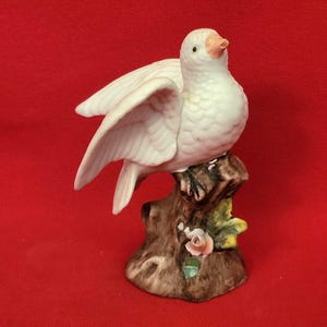 Könnte beinhalten: Eine weiße Keramik-Taubenfigur mit ausgebreiteten Flügeln, die auf einem braunen Baumstumpf sitzt. Die Taube hat einen pfirsichfarbenen Schnabel und ist mit einer kleinen rosa Rose und grünen Blättern verziert.
