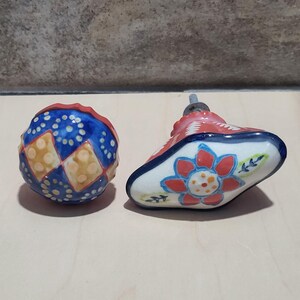 May include: Two decorative ceramic cabinet knobs. One is a blue sphere with white dots and yellow squares. The other is a white, red, and blue floral design. Both have metal screws for installation.
