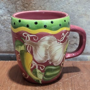 May include: A ceramic mug with a burgundy body, a green and yellow rim, and a burgundy handle. The mug is decorated with illustrations of garlic, peppers, and leaves. Black polka dots adorn the green rim. The mug sits on a wooden surface.