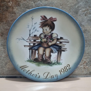 Può includere: Piatto decorativo con bordo blu raffigurante una bambina seduta su una panchina, con un cestino di fiori. Il piatto è iscritto con "Mother's Day 1982" in caratteri dorati. L'opera d'arte ha uno stile vintage dipinto a mano.