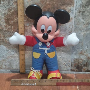 May include: A Mickey Mouse plush doll with black ears, a red shirt, blue overalls, and yellow accents. The doll has white gloves and red shoes with yellow laces. The doll's arms are outstretched, and it has a smiling expression.
