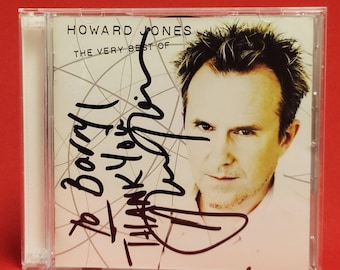 Puede incluir: Una caja de CD con el álbum "The Very Best Of Howard Jones". La portada incluye una foto de Howard Jones y texto manuscrito. La caja es transparente, con un fondo rojo.
