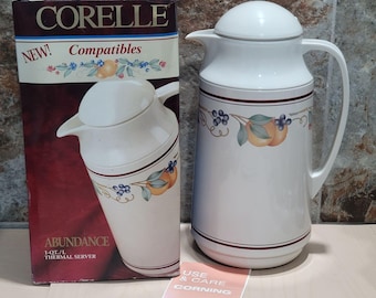 Op de afbeelding: Een witte Corelle Abundance thermoskan met bijpassende deksel, handvat en fruitontwerp. De server staat naast de originele doos, die ook het Corelle-logo en de woorden "NEW! Compatibles" bevat. Er is ook een "Use & Care"-kaart aanwezig.