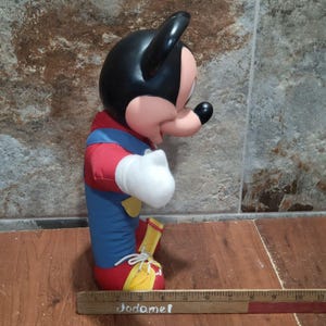 May include: A Mickey Mouse plush toy, featuring a black head and ears, a pink face, and a red, blue, and yellow outfit. The toy is in a seated position, with white gloves and yellow shoes. The word "Jodamel" is written on a wooden ruler.