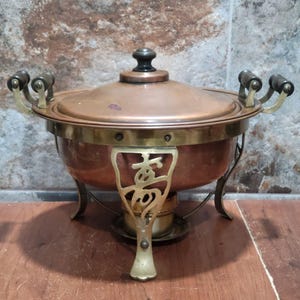Peut inclure: Un plat à réchaud en cuivre avec couvercle, doté de poignées en laiton et d'un support en laiton avec un motif découpé orné. Le couvercle possède un bouton sombre et la base inclut un brûleur central. Les surfaces métalliques ont un fini chaud.