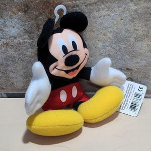 K&ouml;nnte beinhalten: Ein Mickey Mouse Pl&uuml;schtier mit schwarzem Kopf und Ohren, einem beigen Gesicht, wei&szlig;en Handschuhen, roten Shorts mit wei&szlig;en Kn&ouml;pfen und gelben Schuhen. Ein wei&szlig;es Kunststoff-H&auml;ngeetikett ist angebracht.