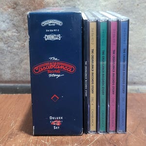 Könnte beinhalten: Ein Deluxe-CD-Set mit dem Titel "The Casablanca Records Story". Die marineblaue Box zeigt das Casablanca Records-Logo und den Text "Chronicles". Das Set enthält vier CDs mit farbigen Rücken, die jeweils mit einer Disc-Nummer und dem Titel Casablanca Records Story beschriftet sind.