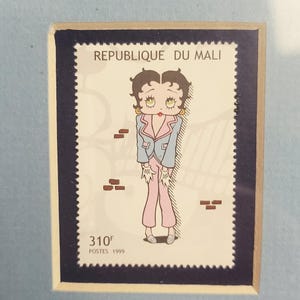 Op de afbeelding: Een postzegel uit de Republiek Mali met een stripfiguur in een roze en blauwe outfit. De postzegel heeft de tekst "REPUBLIQUE DU MALI" bovenaan en "310F POSTES 1999" onderaan. De postzegel is omlijst door een donkerblauwe rand.