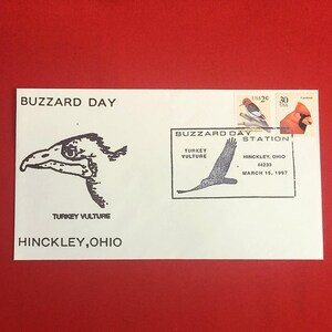 以下が含まれることがあります： 「BUZZARD DAY」と「TURKEY VULTURE HINCKLEY, OHIO」の文字が黒で印刷された白い封筒。ハゲワシのイラストと2枚の切手が特徴です。封筒は赤い背景にあります。