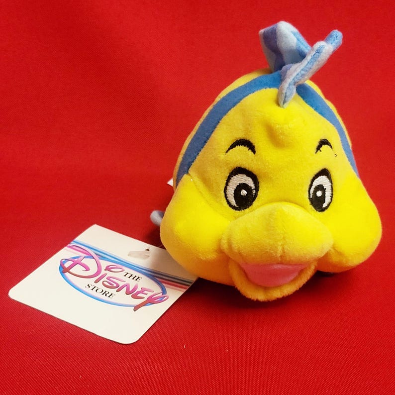 K&ouml;nnte beinhalten: Ein gelbes Pl&uuml;schfisch-Spielzeug mit blauen Akzenten, gro&szlig;en Augen und einem l&auml;chelnden Mund. Das Spielzeug hat ein Etikett mit der Aufschrift "The Disney Store". Der Hintergrund ist einfarbig rot.