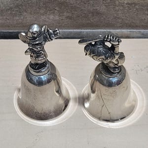 Pu&ograve; includere: Due campane color argento con figurine di personaggi dei cartoni animati sulla parte superiore. Una campana presenta un personaggio con le braccia tese, l'altra ha un personaggio che tiene una mazza da baseball. Le campane hanno una base svasata e poggiano su basi di plastica trasparente.
