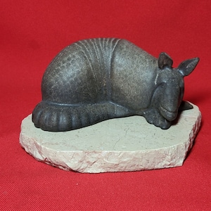 May include: A small, gray, ceramic sculpture of an armadillo curled up sleeping on a beige stone base.