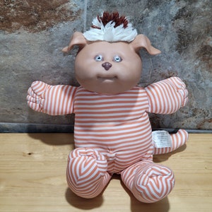 Könnte beinhalten: Ein Plüschtierhund mit braunem und weißem Fell, das einen orange-weiß gestreiften Strampler trägt. Das Spielzeug hat große blaue Augen und eine braune Nase.