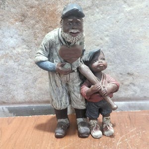 Pode incluir: Uma estatueta de cerâmica que retrata um jogador de beisebol e um menino. A figura mais velha usa um uniforme de beisebol e segura um objeto em forma de coração. O menino segura um bastão de beisebol.