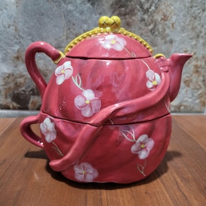 May include: A red ceramic teapot with a floral design and a yellow lid. The teapot is shaped like a flower and has a handle on the side. The teapot is sitting on a wooden surface.