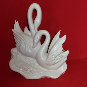 以下が含まれることがあります： 2羽の白鳥が絡み合った白い陶器製の白鳥のプランター。白鳥はスイレンのある池の中にいる。