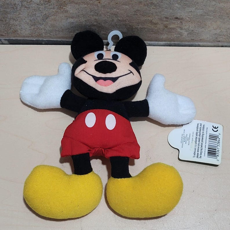 K&ouml;nnte beinhalten: Ein Mickey Mouse Pl&uuml;schtier mit schwarzem Kopf und Ohren, wei&szlig;en Handschuhen, roten Shorts und gelben Schuhen. Das Spielzeug hat ein l&auml;chelndes Gesicht und ausgestreckte Arme. Ein wei&szlig;es Etikett ist angebracht.